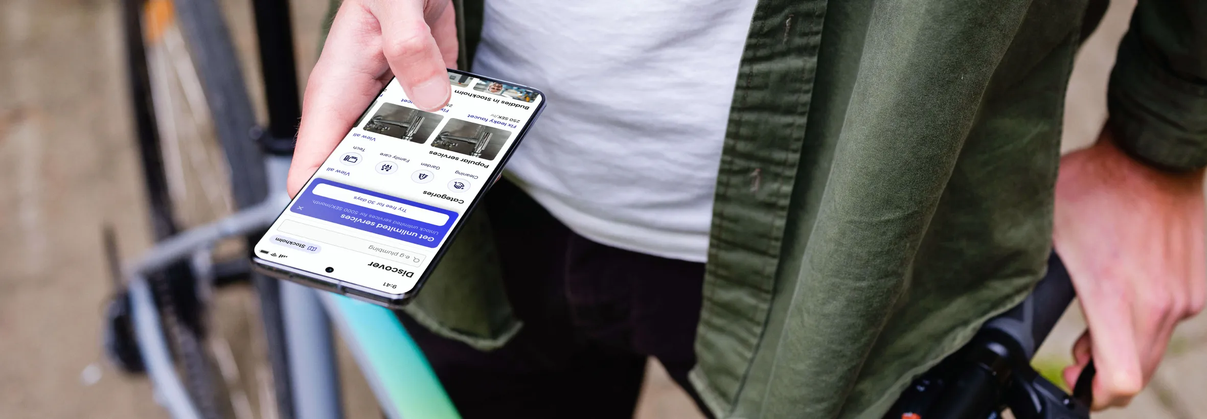 En person i grön jacka håller upp en mobil med Fixat-appen öppen på tjänsteutbudet, samtidigt som hen leder en cykel.