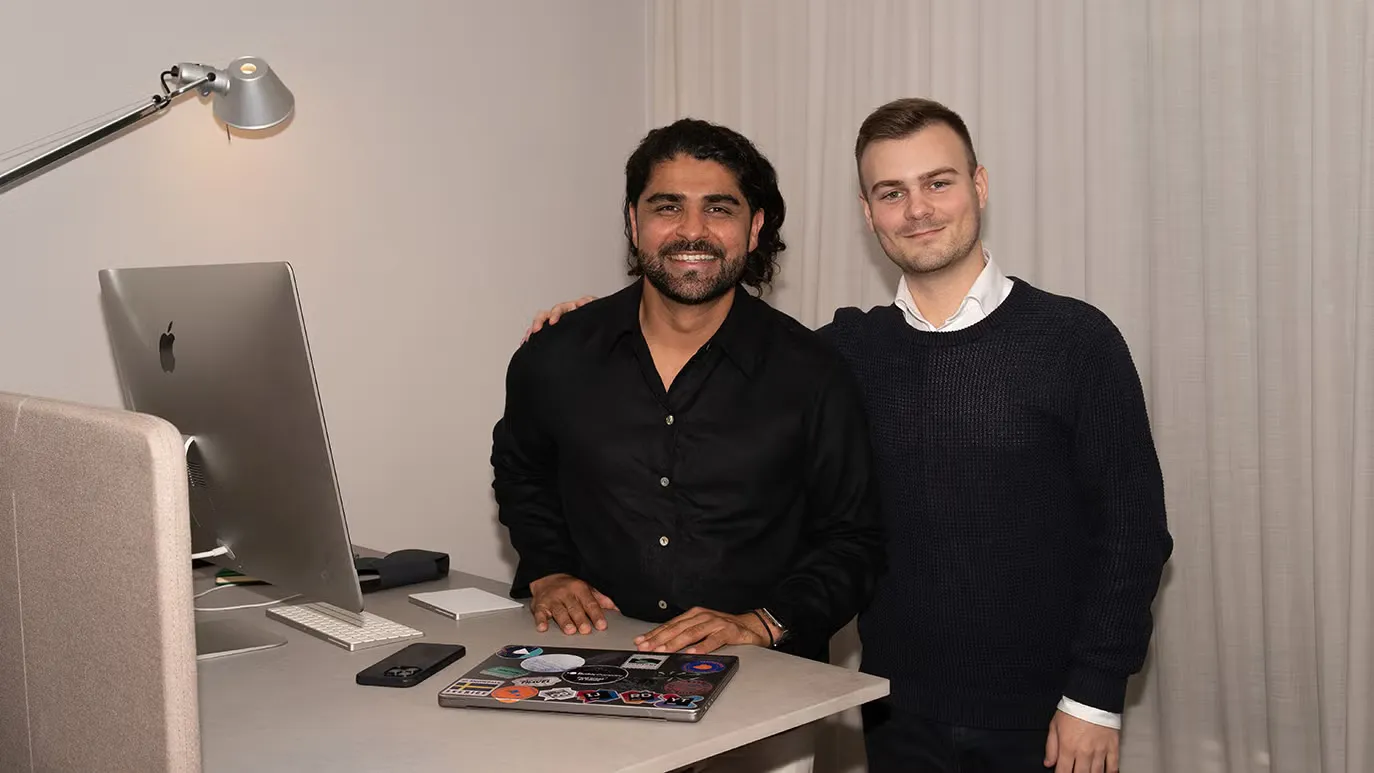 Tahero Nori och en kollega står leende vid ett skrivbord med en stationär dator och en bärbar dator.